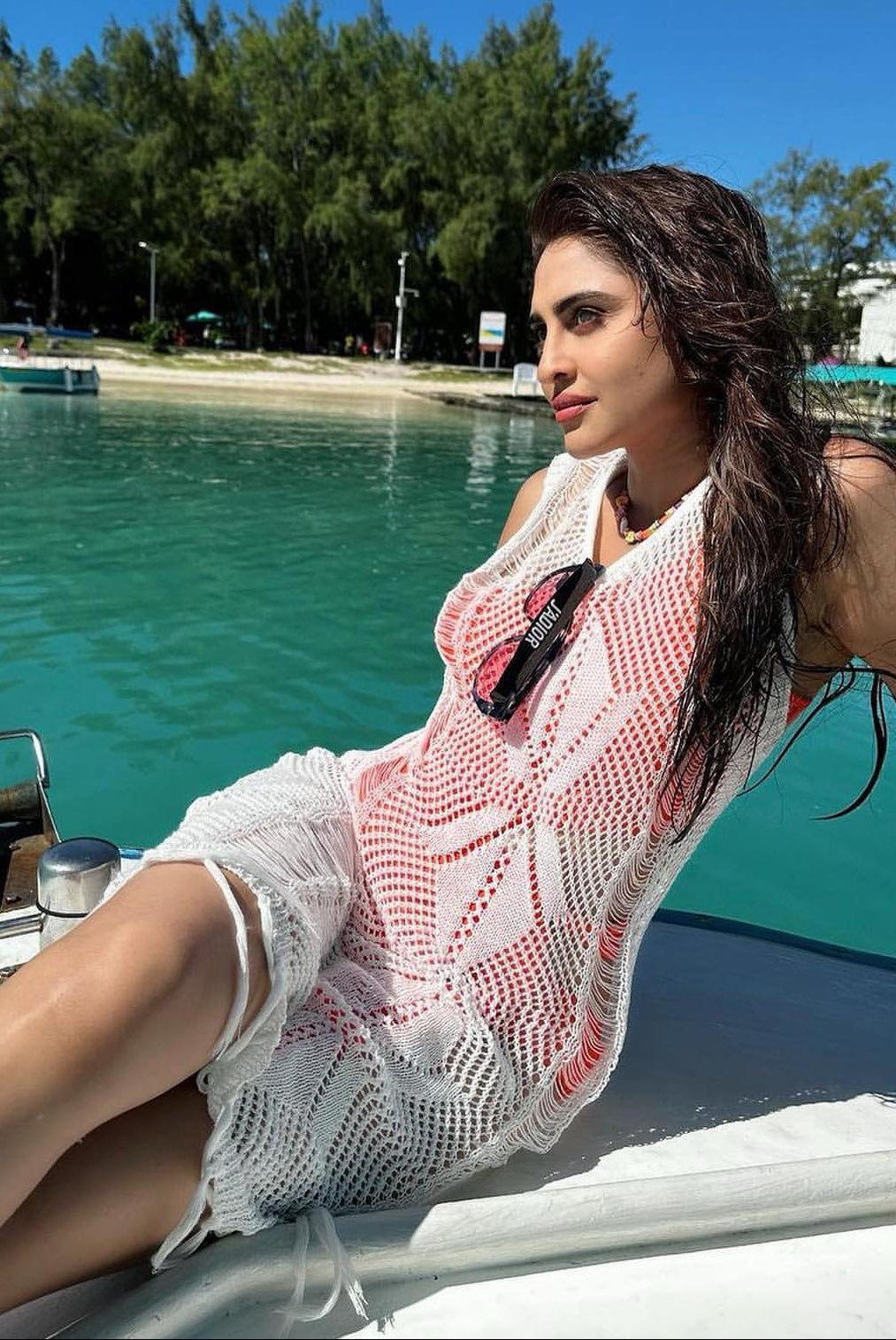 A woman reclining on the bow of a boat in turquoise water, wearing a white crochet halter dress with pink/red dotted trim and sunglasses hanging from the neckline.