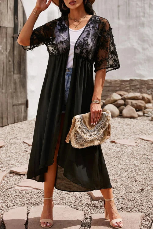 Woman in a black dress with sheer sleeves holding a beaded handbag on a rocky beach.
