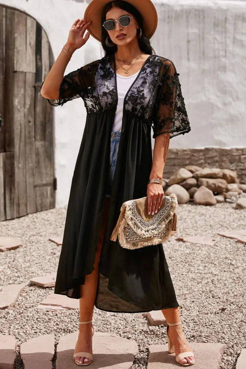 Woman in black floral kimono-style dress with white top and denim shorts, holding a beige handbag, standing outdoors.